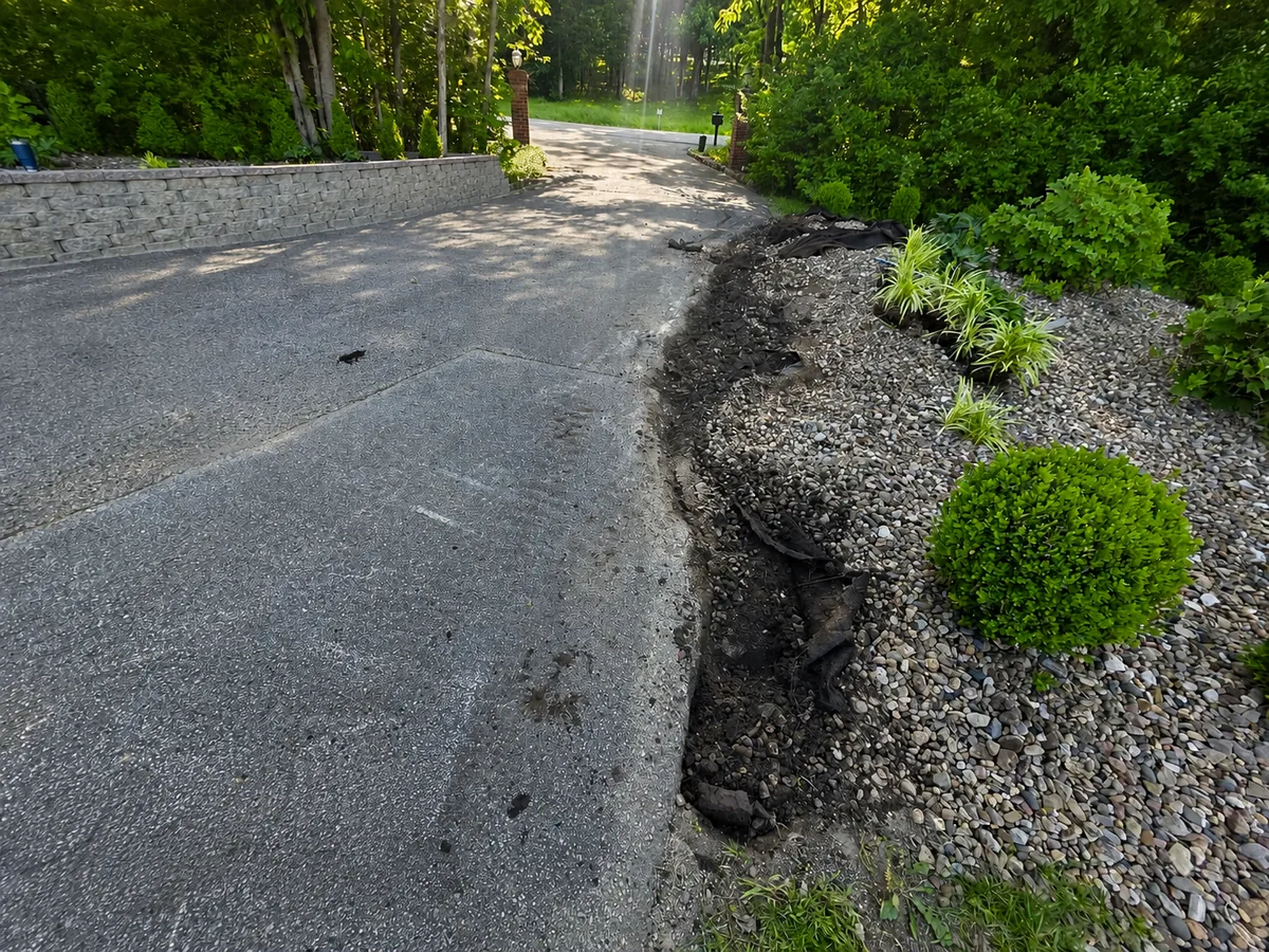 Before photo of a driveway edge with torn fabric, loose rock, and messy bed material
